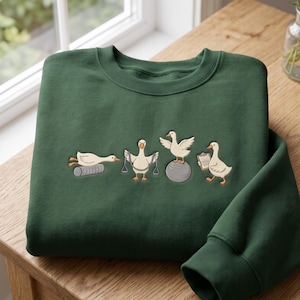 May include: A dark green crewneck sweatshirt featuring an embroidered graphic of four white geese exercising. One goose lies on a foam roller, one holds resistance bands, one stands on a yoga ball, and one holds a clipboard.