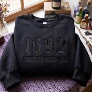 Könnte beinhalten: Ein schwarzes Sweatshirt mit Rundhalsausschnitt und dem gestickten Text 1692 sowie dem Schriftzug THEY MISSED ONE in einer gotischen Schriftart. Das Kleidungsstück hat lange Ärmel mit gerippten Bündchen und einen gerippten Halsausschnitt.