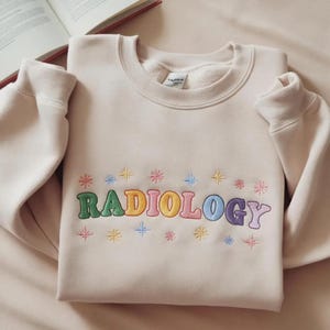 Peut inclure: Un sweat-shirt à col rond beige avec le mot RADIOLOGY brodé en lettres style bulle multicolores sur la poitrine. Le motif est agrémenté de petites formes d'étoiles brodées et colorées au-dessus et au-dessous du texte.