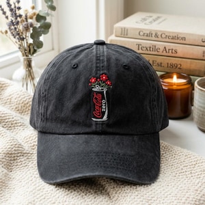 May include: A charcoal black baseball cap featuring an embroidered graphic of a Coca-Cola Zero soda can with red flowers emerging from the top. The cap has a curved brim, a button on the crown, and stitched eyelets for ventilation.