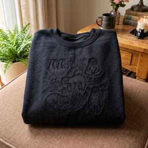 Op de afbeelding: Een zwart sweatshirt met ronde hals voorzien van ton-sur-ton zwart borduurwerk van meerdere luiaards in diverse houdingen, waaronder hangend aan takken en zittend, omringd door gedetailleerde botanische bladeren en bloemmotieven.