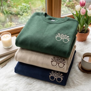 May include: A stack of three crewneck sweatshirts in dark green, beige, and navy blue. Each sweatshirt features an embroidered design on the upper left chest depicting a pair of glasses with a bouquet of various flowers growing out of the frames.