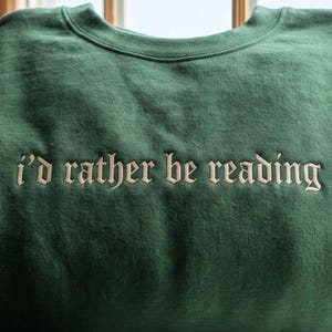 May include: A forest green crewneck sweatshirt featuring the text "i'd rather be reading" embroidered in a light beige, serif-style font across the chest. The garment has a ribbed neckline and a smooth, solid-colored fabric construction.