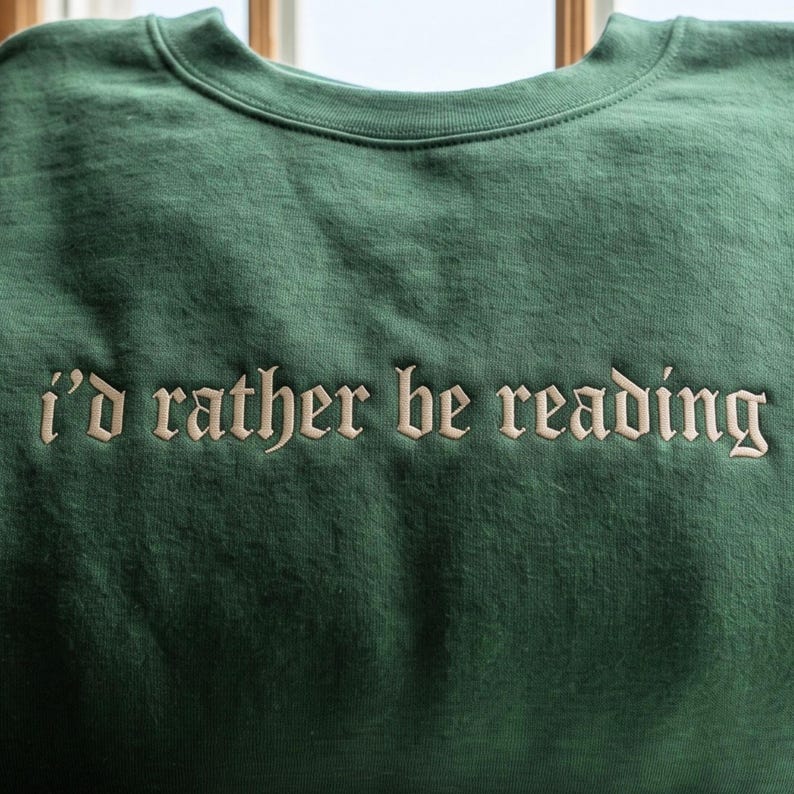 May include: A forest green crewneck sweatshirt featuring the text "i'd rather be reading" embroidered in a light beige, serif-style font across the chest. The garment has a ribbed neckline and a smooth, solid-colored fabric construction.