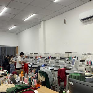 May include: A row of industrial embroidery machines with multiple needles and thread spools arranged in a bright workshop. Several people are operating the machinery, which is set up on white tables. Various colorful garments and fabric items are placed near the equipment.