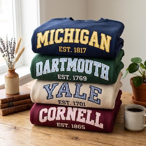 May include: A stack of four crewneck sweatshirts in navy, dark green, beige, and maroon. Each features university names and establishment years in a gingham-patterned applique: MICHIGAN EST. 1817, DARTMOUTH EST. 1769, YALE EST. 1701, and CORNELL EST. 1865.