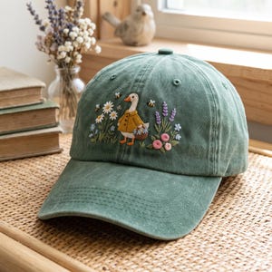 May include: A sage green baseball cap featuring an embroidered design of a white goose wearing a yellow coat and carrying a basket. The embroidery includes colorful flowers, green leaves, and small bees surrounding the goose on the front panel.
