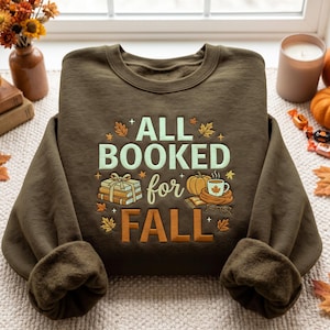 Peut inclure: Un sweat-shirt à col rond vert olive foncé avec le texte ALL BOOKED for FALL en vert clair et orange. Le motif comprend une pile de trois livres attachés par un ruban, une citrouille, une tasse fumante et des lunettes, entourés de feuilles d'érable.