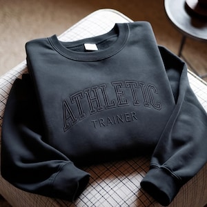 May include: A charcoal gray crewneck sweatshirt featuring the word ATHLETIC in a large, arched, outlined font above the word TRAINER in a smaller, solid, sans-serif font. The garment has long sleeves with ribbed cuffs and a ribbed neckline.