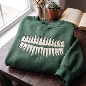Op de afbeelding: Een bosgroene sweater met ronde hals en een grote, gecentreerde borduursel van een volledig menselijk gebit in gebroken wit garen. Het ontwerp toont zowel de bovenste als de onderste rij tanden, inclusief kiezen en snijtanden.