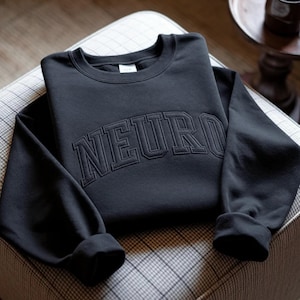 以下が含まれることがあります： 胸元に黒い糸でNEUROという文字が太字のアーチ状カレッジフォントで刺繍された、黒いクルーネックのスウェットシャツ。長袖で、袖口と襟元はリブ編みになっています。