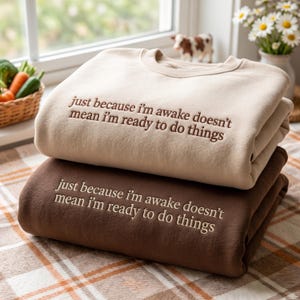 May include: Two crewneck sweatshirts stacked on top of each other. The top sweatshirt is beige with dark brown embroidered text that reads, just because i'm awake doesn't mean i'm ready to do things. The bottom sweatshirt is dark brown with beige text.