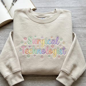May include: A beige crewneck sweatshirt featuring the embroidered text "Surgical Technologist" in a multi-colored pastel script font. The design is accented with small embroidered flowers and starburst shapes in shades of pink, blue, yellow, and green.