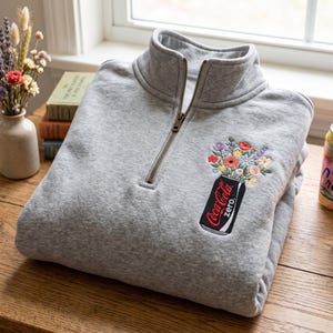 以下が含まれることがあります： 胸元に刺繍が施された、グレーのハーフジップスウェットシャツ。黒いコカ・コーラゼロの缶を花瓶に見立て、赤、青、黄色の刺繍された花々が活けられているデザインが特徴です。