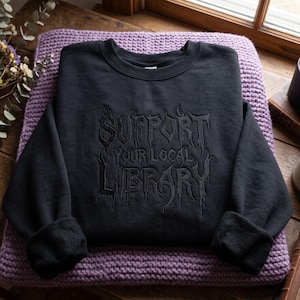 Peut inclure: Un sweat-shirt noir à manches longues et col rond avec une broderie noire sur la poitrine. Le texte brodé indique "SUPPORT YOUR LOCAL LIBRARY" dans une police gothique stylisée et dentelée avec des détails en forme de gouttes.