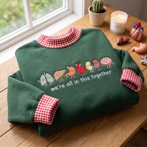 May include: A dark green sweatshirt with a red and white gingham collar and cuffs. The front features an embroidered graphic of cartoon human organs holding hands with the white text "we're all in this together" printed underneath.