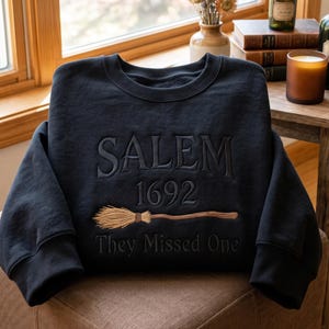 Könnte beinhalten: Ein schwarzes Sweatshirt mit Rundhalsausschnitt und gesticktem Text SALEM 1692 sowie They Missed One, ergänzt durch eine gestickte Grafik eines braunen Besens mit beigen Borsten, die mittig auf der Vorderseite platziert ist.