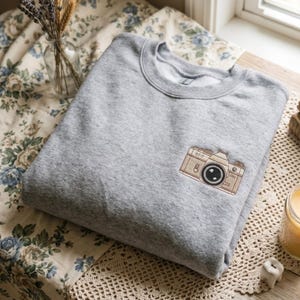May include: A heather gray crewneck sweatshirt featuring a small, detailed embroidery of a vintage-style camera on the left chest. The camera is rendered in shades of tan and brown with black accents and the text S7-7 visible on its top surface.