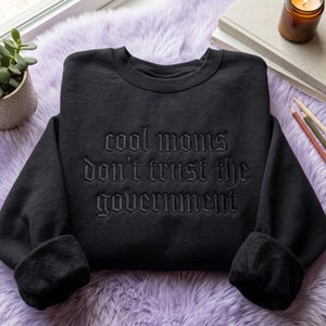 May include: A black long-sleeve crewneck sweatshirt featuring the embroidered text "cool moms don't trust the government" in a matching black gothic-style font across the center of the chest. The garment has a ribbed collar and cuffs.