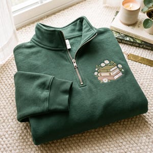 May include: A dark forest green quarter-zip sweatshirt featuring a small embroidered graphic on the left chest. The embroidery depicts a stack of three books in muted tones surrounded by white daisy flowers with yellow centres.