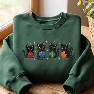 May include: A dark green crewneck sweatshirt featuring an embroidered graphic of four black cats, each holding a different colored polyhedral gaming die. The cats are surrounded by small gold star accents, creating a whimsical and detailed design.