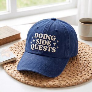 May include: A navy blue baseball cap featuring the embroidered white text DOING SIDE QUESTS in a bold, serif font, accented by small four-pointed star symbols on either side of the text, with a structured crown and a curved brim.