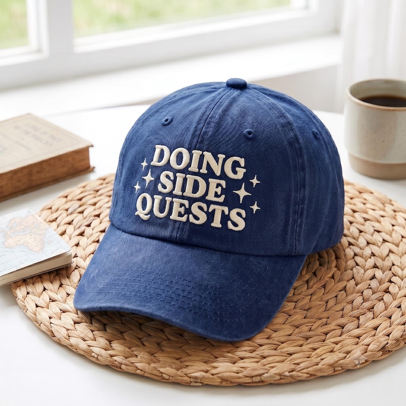 May include: A navy blue baseball cap featuring the embroidered white text DOING SIDE QUESTS in a bold, serif font, accented by small four-pointed star symbols on either side of the text, with a structured crown and a curved brim.