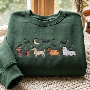May include: A dark forest green crewneck sweatshirt featuring an embroidered design of four dachshunds in Halloween costumes: a witch, a mummy, a hot dog, and a ghost. The scene includes a crescent moon, small stars, and black bats above the dogs.