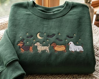 May include: A dark forest green crewneck sweatshirt featuring an embroidered design of four dachshunds in Halloween costumes: a witch, a mummy, a hot dog, and a ghost. The scene includes a crescent moon, small stars, and black bats above the dogs.
