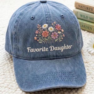 K&ouml;nnte beinhalten: Eine blaue Jeans-Baseballkappe mit dem gestickten Text "Favorite Daughter" in Wei&szlig;. Die Kappe hat ein Blumenmuster mit rosa, wei&szlig;en und orangefarbenen Blumen. Der Hut hat einen gebogenen Schirm und einen Knopf oben.