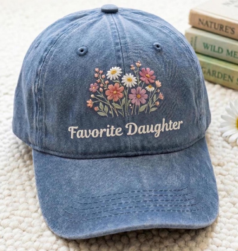 K&ouml;nnte beinhalten: Eine blaue Jeans-Baseballkappe mit dem gestickten Text "Favorite Daughter" in Wei&szlig;. Die Kappe hat ein Blumenmuster mit rosa, wei&szlig;en und orangefarbenen Blumen. Der Hut hat einen gebogenen Schirm und einen Knopf oben.