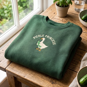 Könnte beinhalten: Ein dunkelgrüner Rundhals-Pullover mit einer Stickerei, die eine weiße Gans mit einer grünen Gewürzgurke zeigt. Der Schriftzug "PICKLE PRINCESS" ist in weißen Buchstaben bogenförmig über der Gans angebracht. Der Pullover hat gerippte Bündchen.