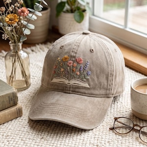 May include: A tan baseball cap featuring an embroidered design of an open book with colourful wildflowers growing out of the pages. The cap has a structured crown, a curved brim with visible stitching, and a button at the top centre.