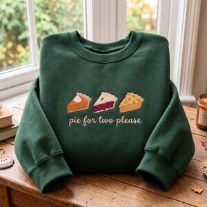 May include: A dark forest green crewneck sweatshirt featuring an embroidered design of three distinct pie slices and the text "pie for two please" in a light cream script font centred on the chest area of the garment.