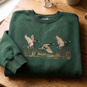 May include: A dark forest green crewneck sweatshirt featuring an embroidered design of three mallard ducks in flight over a marshy pond with cattails. The embroidery uses shades of brown, white, green, and tan to depict the ducks and reeds.