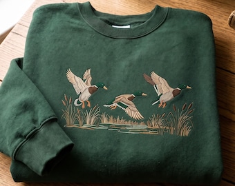 May include: A dark forest green crewneck sweatshirt featuring an embroidered design of three mallard ducks in flight over a marshy pond with cattails. The embroidery uses shades of brown, white, green, and tan to depict the ducks and reeds.