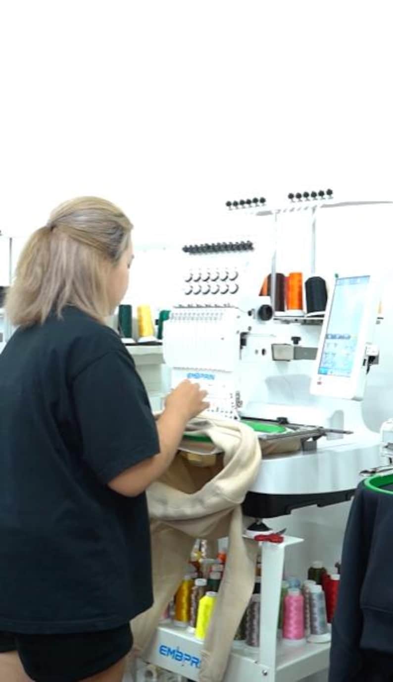 May include: A white industrial embroidery machine with multiple thread spools in various colors including orange, black, and green. The machine features a digital screen and is mounted on a white rolling cart filled with numerous spools of thread.