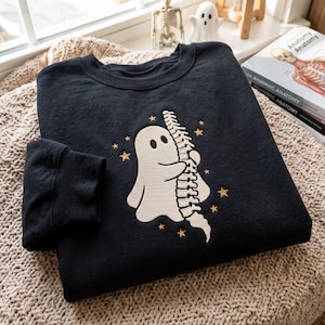 May include: A black crewneck sweatshirt featuring an embroidered design of a cream-coloured ghost hugging a human spine. The graphic is surrounded by small, scattered gold-coloured stars. The sweatshirt has long sleeves with ribbed cuffs.