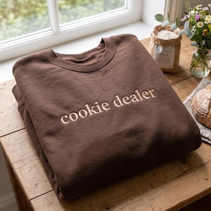 May include: A dark brown crewneck sweatshirt featuring the text "cookie dealer" embroidered in a light tan thread across the chest. The garment has a ribbed neckline and long sleeves, showcasing a simple design with clean, centred lettering.