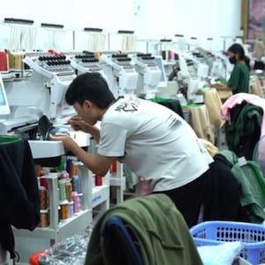 May include: A row of white industrial embroidery machines with multiple thread spools in a workshop setting. A person in a white t-shirt operates one machine, while another person works at a station further down the line with various garments.
