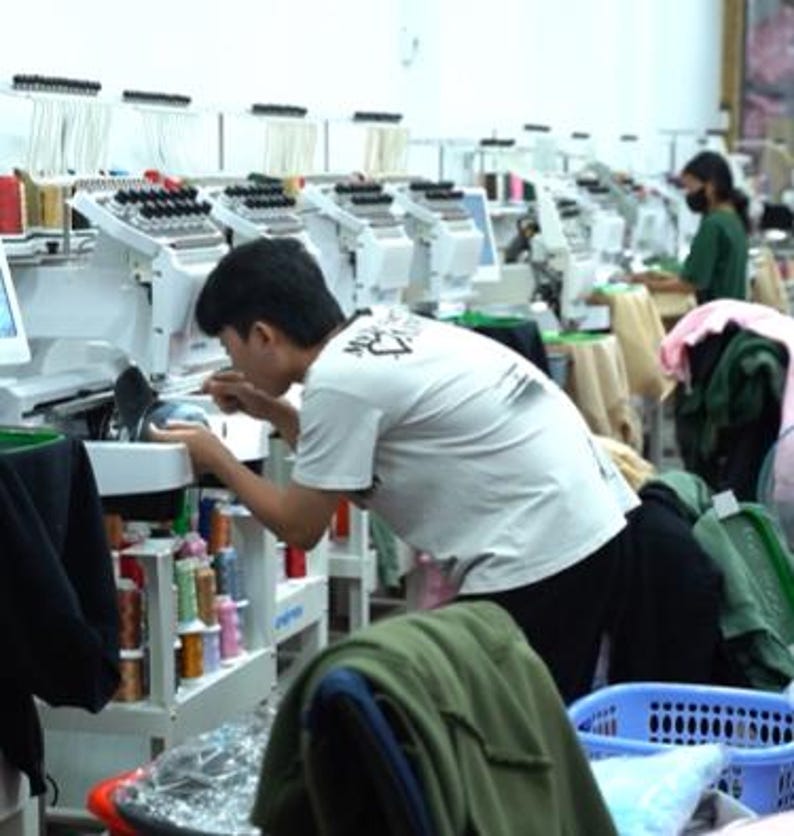 May include: A row of white industrial embroidery machines with multiple thread spools in a workshop setting. A person in a white t-shirt operates one machine, while another person works at a station further down the line with various garments.