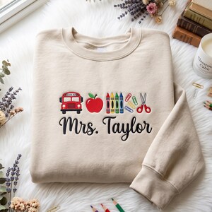 May include: A beige crewneck sweatshirt featuring embroidered school-themed icons including a red school bus, a red apple, colourful crayons, paper clips, and red scissors. Below the icons, the name Mrs. Taylor is embroidered in black script.