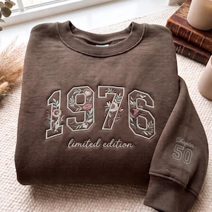 May include: A dark brown crewneck sweatshirt featuring the embroidered text 1976 with floral accents inside the numbers. Below the numbers is the text limited edition. The sleeve features the embroidered text Chapter 50 in a light thread.