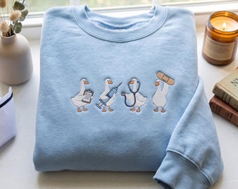May include: A light blue crewneck sweatshirt featuring an embroidered design of four white geese dressed as medical professionals. One goose holds a clipboard, another a syringe, one wears a stethoscope, and the last has a bandage on its head.
