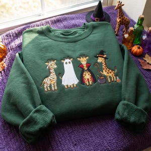 May include: A dark green crewneck sweatshirt featuring an embroidered graphic of four giraffes in Halloween costumes. The giraffes are dressed as a mummy, a ghost, a vampire, and a witch with a cauldron, all arranged in a horizontal line across the chest.