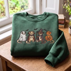 Op de afbeelding: Een donkergroene sweater met ronde hals en een grafische print van vier gekko's in Halloween-kostuums. Eén gekko draagt een wit spooklaken, één een vampiercape, één een heksenhoed met bezem en één zit in een uitgesneden oranje pompoen.