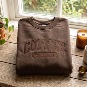 May include: A dark brown crewneck sweatshirt featuring the word COFFEE in a large, arched, embroidered font, with the word WEATHER centred below it inside a rectangular embroidered border. The garment has a ribbed collar and cuffs.
