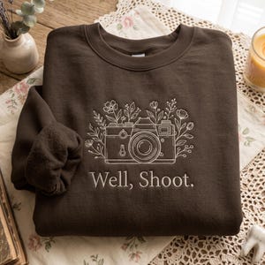 Peut inclure: Un sweat-shirt à col rond marron foncé avec une broderie au trait blanc représentant un appareil photo vintage entouré de fleurs sauvages. Le texte "Well, Shoot." est brodé en blanc sous le motif de l'appareil photo sur le devant.