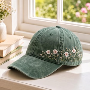 May include: A sage green baseball cap featuring an embroidered design of delicate white and pink daisy flowers with green stems and leaves wrapping around the side of the crown. The cap has a structured brim and a classic six-panel construction.