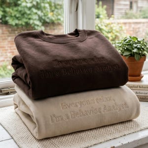 May include: Two folded crewneck sweatshirts stacked on top of each other. The top sweatshirt is dark brown, and the bottom one is beige. Both feature the embroidered text, "Everyone relax, I'm a Behavior Analyst." in a matching thread color.